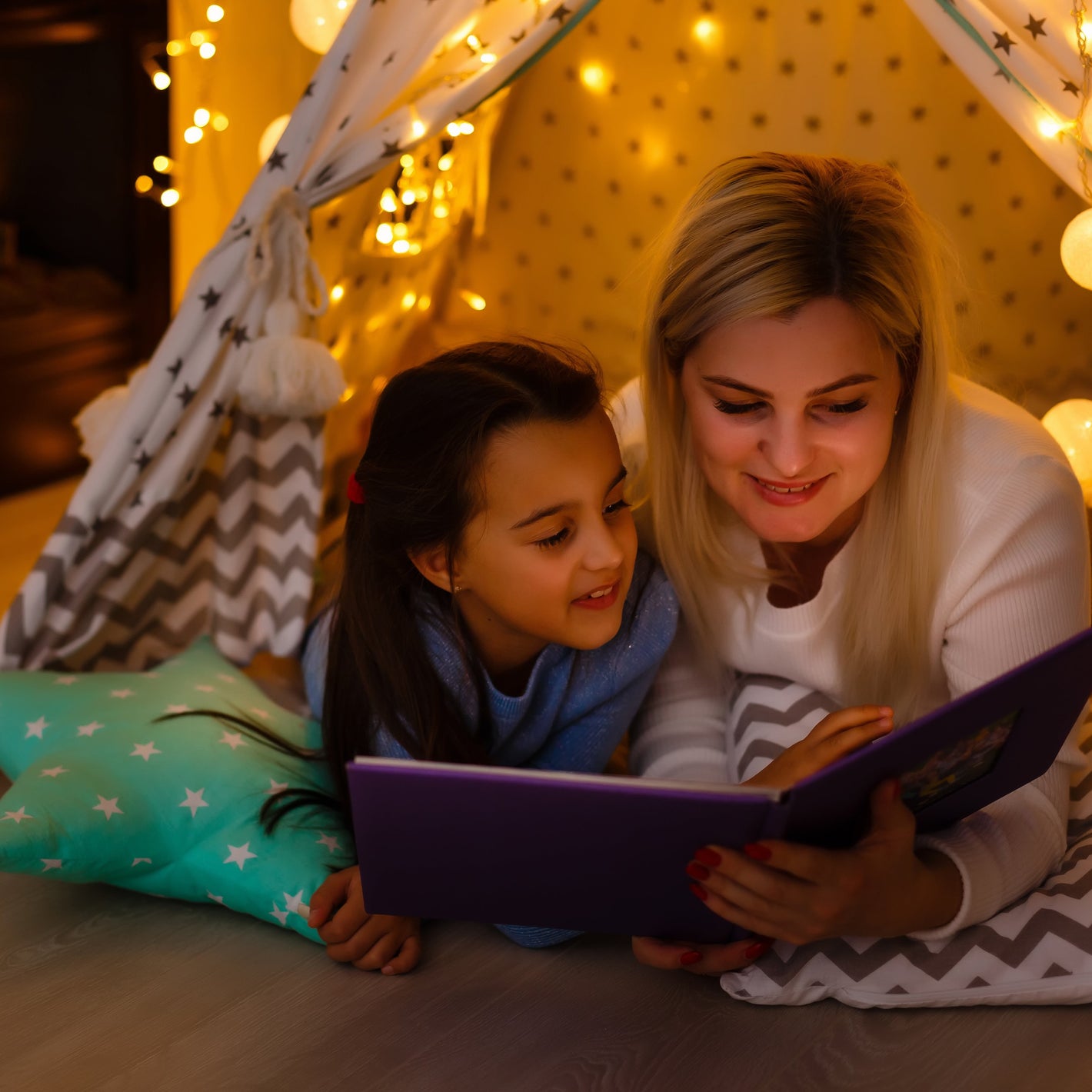 Woman and child reading a book together in a cozy indoor setting with lights and a fireplace.